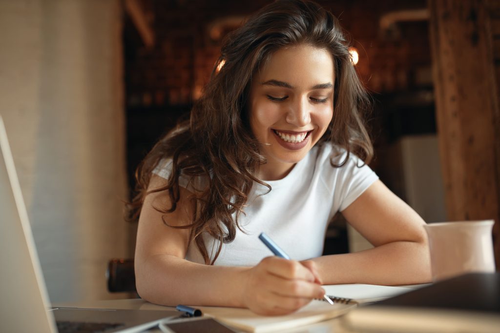 Fotografia de mulher sorrindo e escrevendo em um caderno.